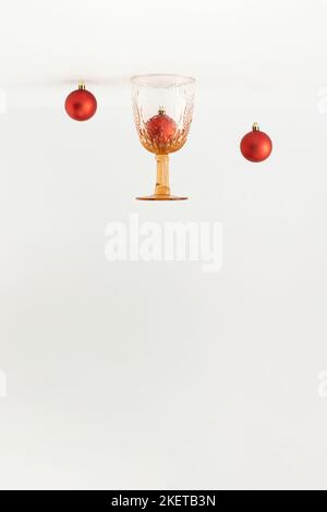Vintage drinking glass and red Christmas baubles decor turned upside down on white background. Minimal New Year concept. Stock Photo