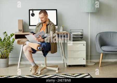 Full length portrait of man with prosthetic leg reading document at workplace in office, copy space Stock Photo