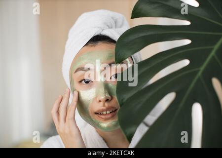 Young healthy asian woman with green cosmetic mask on face and towel on head hiding her face behind monstera leaves. Beautiful female doing skin care procedures in exotic home interior. Stock Photo