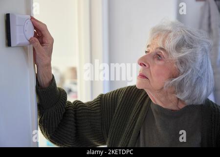 Worried Senior Woman Turning Down Central Heating Thermostat At Home In Energy Crisis Stock Photo