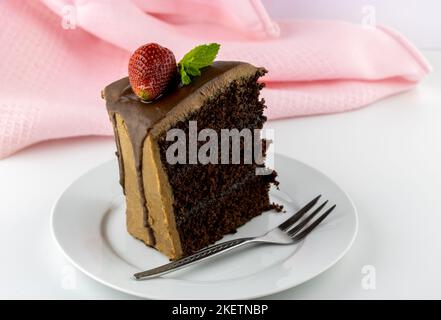 Slice of chocolate layer cake with fudge frosting and strawberry on white with pink in background Stock Photo