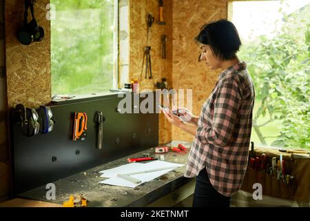 Female business owner using digital tablet in workshop, industrial woman engineer working with blueprints and tablet in the workshop. Stock Photo