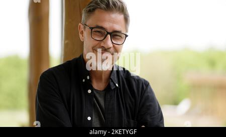 Portrait of happy casual middle age man in glasses standing on balcony at home relaxing, smiling. Stock Photo