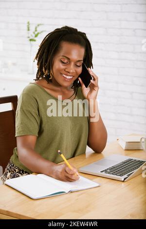 Smililing female freelancer talking on phone and taking notes Stock Photo