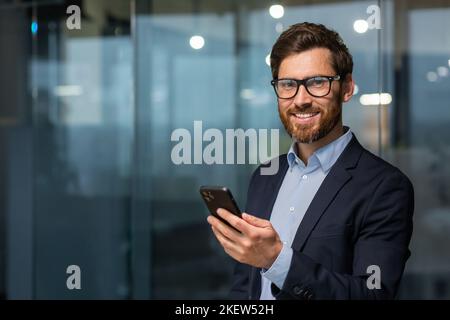 Portrait of successful mature businessman investor, man in glasses and beard smiling and looking at camera, boss in suit at work inside office holding smartphone. Stock Photo