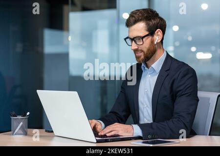 Successful mature businessman at work with laptop inside office, man in business suit sitting at desk typing on computer, satisfied with achievement result and work. Stock Photo