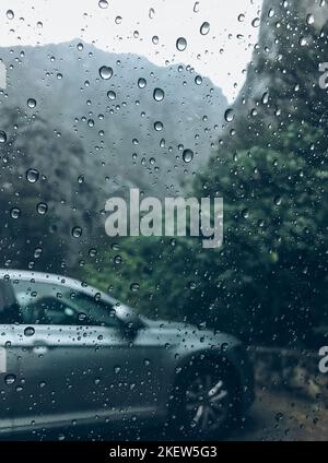 Raindrops on the clean car glass image with silver color car on the background. Deep autumn cold rainy foggy day photo on the mountain parking spot. W Stock Photo