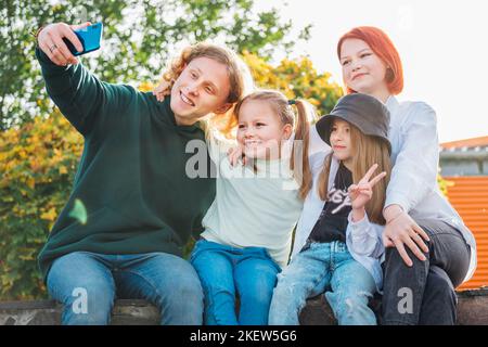 Portraits of three smiling sisters and brother teen taking selfie portrait using a modern smartphone camera. Careless happy young teenhood, childhood Stock Photo