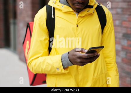Focus on hand of young deliveryman with mobile phone messaging or looking through orders of clients while standing in front of camera Stock Photo