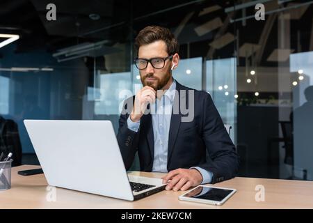 Senior businessman in glasses and beard is thinking about a decision, man is working inside the office and using laptop at work, mature investor in a business suit is sitting at the table Stock Photo