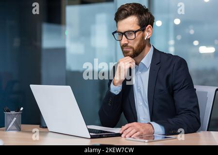 Senior businessman in glasses and beard is thinking about a decision, man is working inside the office and using laptop at work, mature investor in a business suit is sitting at the table Stock Photo
