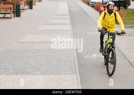 Young African American male courier with backpack riding bicycle while moving along road in park or other urban environment Stock Photo