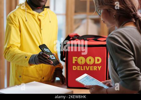 African American male courier in yellow jacket holding smartphone with online menu on screen while showing it to young waitress Stock Photo