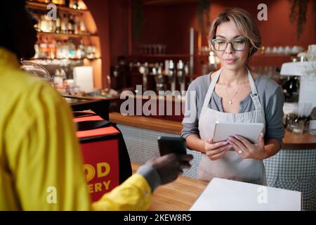 Focus on young waitress with tablet looking at screen of smartphone held by courier while checking online menu and list of online orders Stock Photo