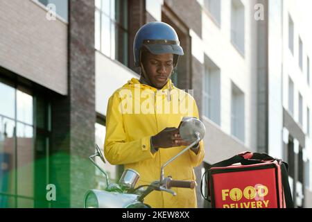 Young African American man in courier uniform and helmet using smartphone while checking data about orders of clients Stock Photo