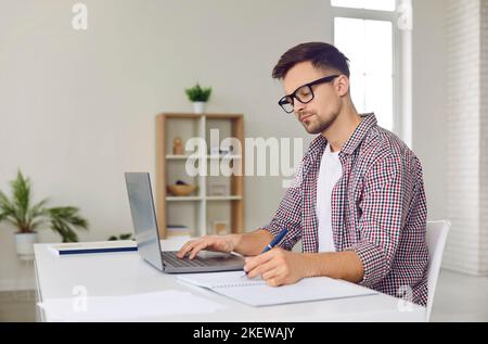 Remote student in glasses sitting at desk with laptop computer and writing in notebook Stock Photo