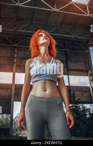Strong female athlete wearing sportswear taking break after exercising. Fit woman standing inside abandoned warehouse. Stock Photo