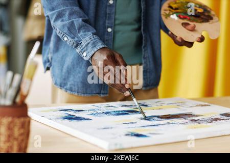 Hand of young African American male artist with color palette painting with acrylic paints on canvas while standing by workplace Stock Photo