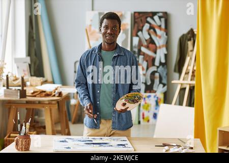 Young successful African American male artist in casualwear holding wooden color palette with acrylic paints while creating artwork Stock Photo