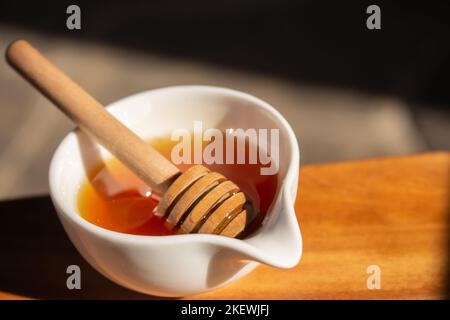 Honey with dipper in small white bowl. Homemade honey with wooden stick in sunlight. Honey in jar. Natural sweetener. Healthy nutrition. Stock Photo