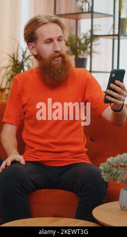 Portrait of caucasian bearded man making smartphone call conversation with friends. Happy excited young guy at home in room on orange couch enjoying mobile phone loudspeaker talking. Vertical view Stock Photo