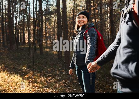Couple holding hands enjoying trip while vacation day. Hikers with backpacks walking on forest path on sunny day. Active leisure time close to nature Stock Photo