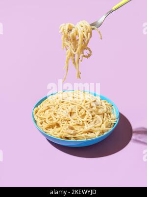 Pasta Cacio e Pepe in a blue bowl isolated on a purple background. Taking spaghetti with a fork from a bowl in bright light against a colorful backgro Stock Photo