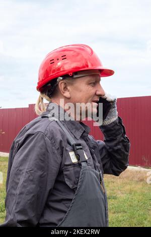 An adult male worker in overalls and a hard hat is talking on the phone on the street. Stock Photo