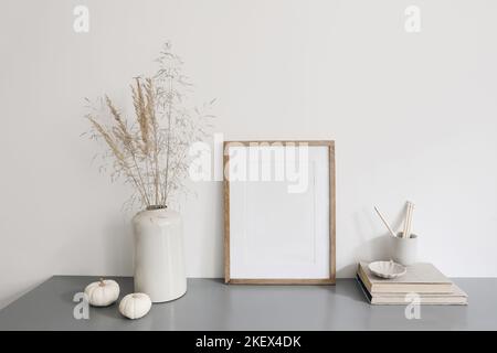 Blank vertical wooden picture frame mockup. Vase with dry grass and white pumpkins on desk. Pencils in ceramic holder, old books. Working space, home Stock Photo