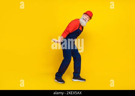 Photo of funky cheerful senior guy dressed uniform overall red hardhat holding arms heavy empty space isolated yellow color background Stock Photo