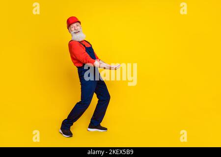 Photo of good mood funky senior guy dressed uniform overall red hardhat holding arms heave empty space isolated yellow color background Stock Photo