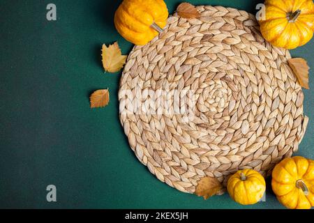 Cozy hygge table. Top view of wicker place mat, pumpkins, autumn leaves on green table. Autumn table setting. Festive Thanksgiving background. Restaur Stock Photo