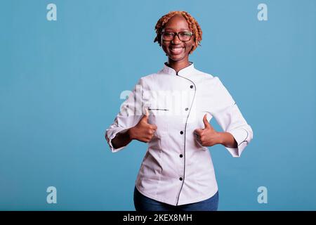Strong and confident african american chef expressing ok symbol to camera. Brave and proud woman wearing kitchen uniform while giving thumbs up sign on blue background. Stock Photo