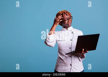 Female chef dressed in work uniform using laptop with confused expression while reading new recipes on restaurant menu. Expert female cook using mobile device in studio shot. Stock Photo