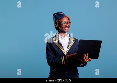 Smiling african american flight attendant wears uniform using laptop during break time at work. Stewardess checking emails from notebook looks happy for good news, studio shot. Stock Photo