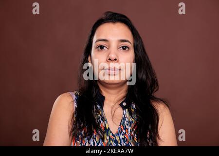 Indian woman looking at camera medium closeup portrait. Calm lady with neutral facial expression close view, relaxed smiling person front view studio medium shot on brown background Stock Photo