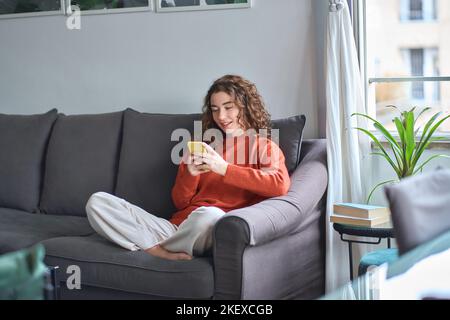 Smiling young woman sitting on sofa using mobile phone at home. Stock Photo
