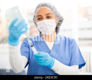 Female nurse wearing a protective mask fills a syringe with saline solution in the treatment room Stock Photo