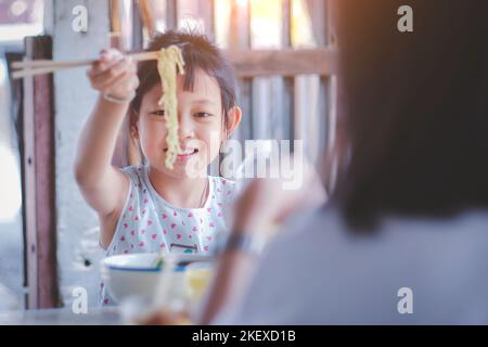 Smiling Asian girls happily eating noodles with their mother sitting together Stock Photo