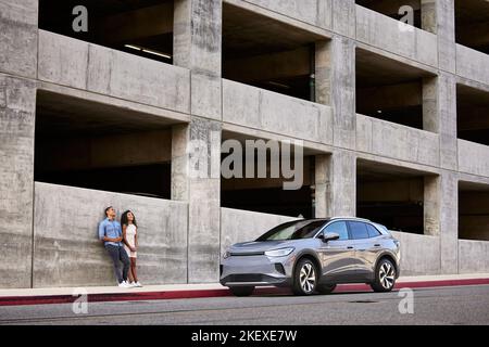Couple leaning on building wall by electric car parked at roadside Stock Photo