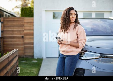 Contemplative woman with smart phone leaning on electric car Stock Photo