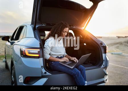Young woman using laptop sitting in car trunk during sunset Stock Photo