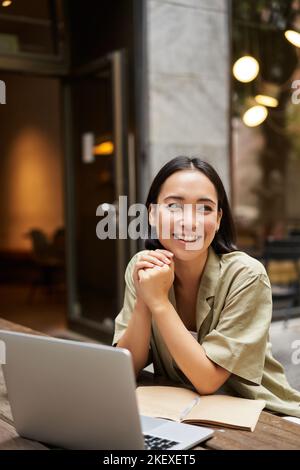 Portrait of asian girl works outdoors in cafe, sits with laptop, studies, smiles happy Stock Photo