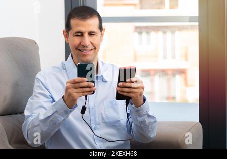 Happy man charging a smartphone with a portable charger sitting on a couch at home Stock Photo