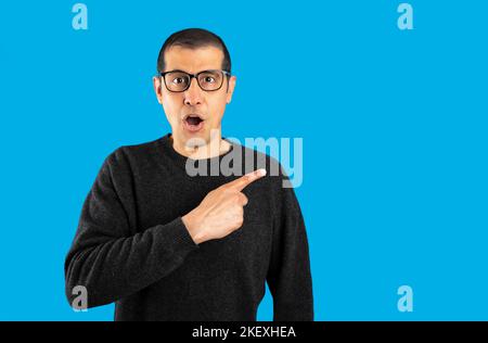 Studio shot of a hispanic man wearing casual sweater and glasses over blue background Surprised pointing with finger to the side, open mouth amazed ex Stock Photo