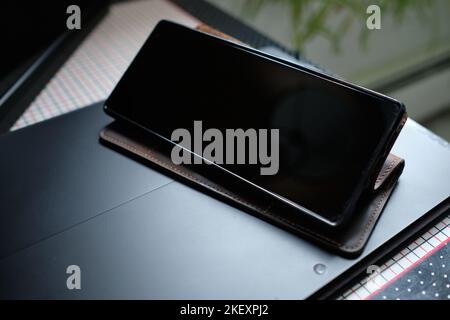 Black cellphone in a brown leather case is stood up on top of a black lap top on a side table at home with the screen turned off Stock Photo