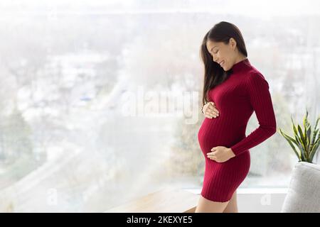 Asian pregnant woman at home. Prenatal pregnancy photo of beautiful model standing holding belly in red dress indoors with winter background and copy Stock Photo