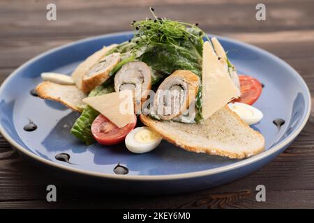 Caesar salad with croutons, quail eggs, cherry tomatoes and grilled chicken roll in a blue plate on a dark wooden table Stock Photo