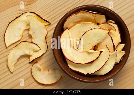 Dried apple slices in a brown wood bowl next to spilled dried apple slices on light wood background. Top view. Stock Photo