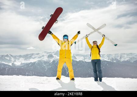 Couple of skier and snowboarder are standing at mountain top, holding ski and snowboard in hands and having fun. Winter sports at ski resort concept Stock Photo